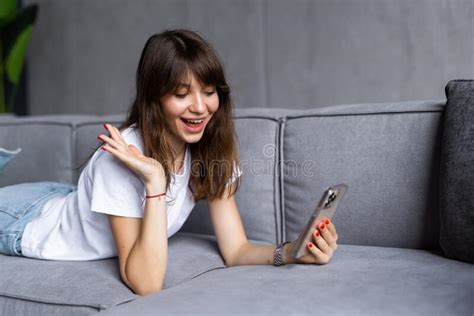 Happy Young Woman With Toothy Smile Waving Hand Looking At Camera Sitting On Couch At Home