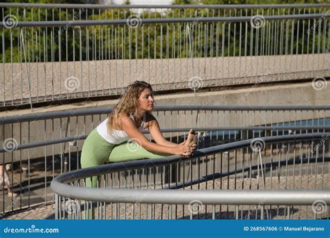 Middle Aged Blonde Woman In Green Leggings And White Top Doing Stretching Exercises On A Railing