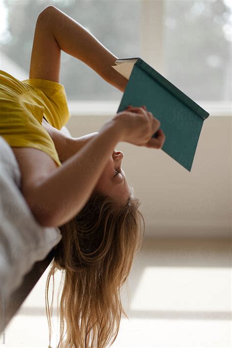 Girl Hanging Upside Down Off Edge Of Bed While Reading A Book By