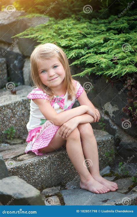 A Little Girl In A Dress Sits On The Stone Steps In The Garden Bare