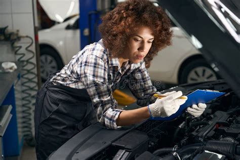 Female Technician In Work Clothes Stands Near Car With Open Hood Stock