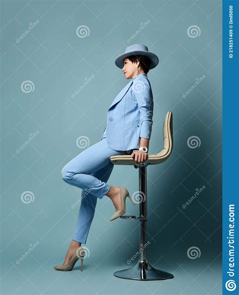 Short Haired Brunette Woman In Blue Business Suit Hat High Heeled Shoes Sitting On Bar Stool