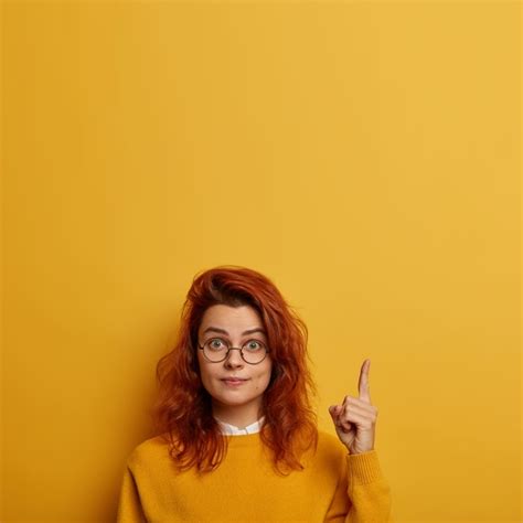 좀 봐 놀란 빨간 머리 유럽 여성은 둥근 안경을 쓰고 위의 포인트는 새로운 제품을 보여주고 복사 공간을 광고합니다 무료 사진