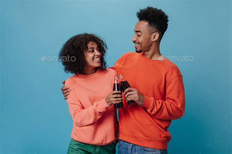 Curly Guy And His Girlfriend Clinking Soda Water Bottles Attractive Brunette Woman In Orange