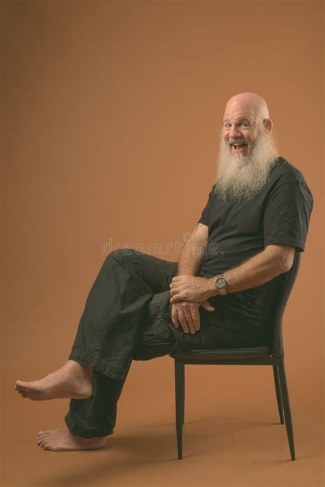 Mature Bald Man With Long Beard Sitting On Chair In Black And White