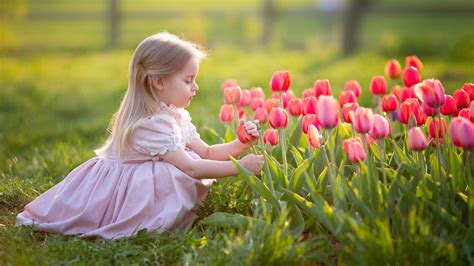 Cute Little Girl Is Touching Tulip Sitting On Grass Wearing Light Pink