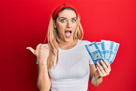 Young Caucasian Woman Holding 1000 Hungarian Forint Banknotes Pointing Thumb Up To The Side