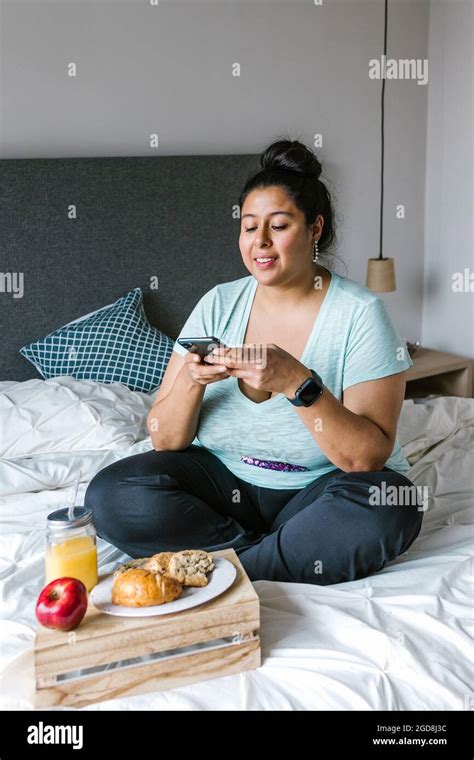 Young Mexican Plus Size Woman Sitting In Bed Using Phone While Having