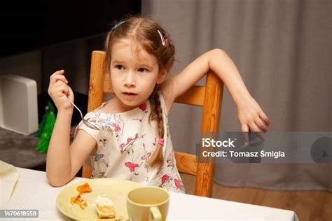 손에 포크를 들고 음식을 먹는 어린 소녀는 조잡하고 부주의한 편안한 자세방식으로 의자에 앉아 있습니다 의자 등받이에 팔을