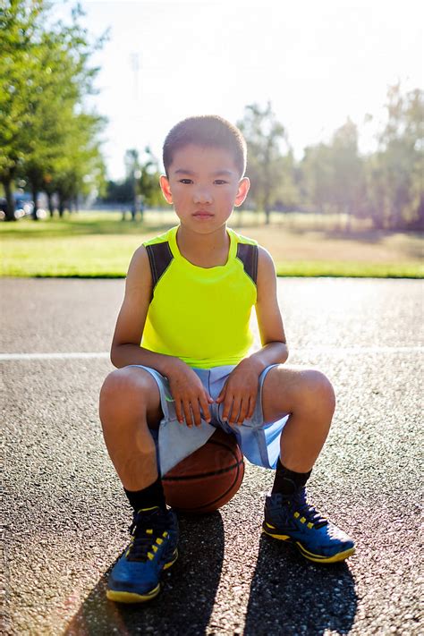 Asian Kid Sitting On A Basketball In An Outdoor Basketball Court By