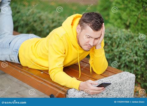 Young Man Lying On Her Stomach Outside On A Bench With Her Legs Up And Browsing Her Cell Phone