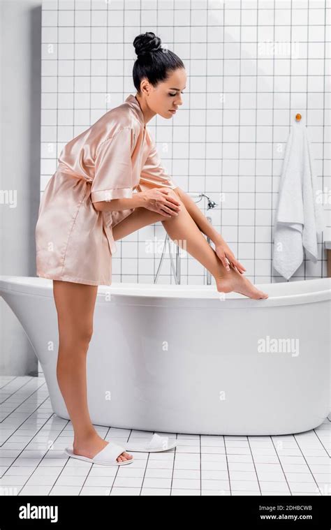 Side View Of Brunette Woman In Satin Bathrobe Touching Leg Near Bathtub In Modern Bathroom Stock