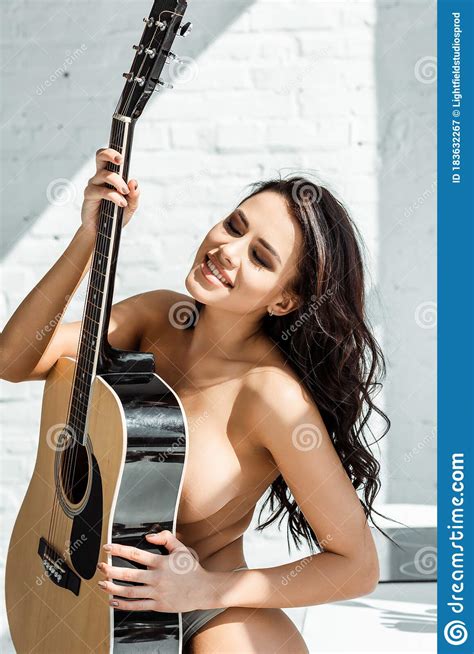 Mujer En Topless Sonriendo Mientras Sostiene Guitarra Acústica En La Cocina Imagen de archivo