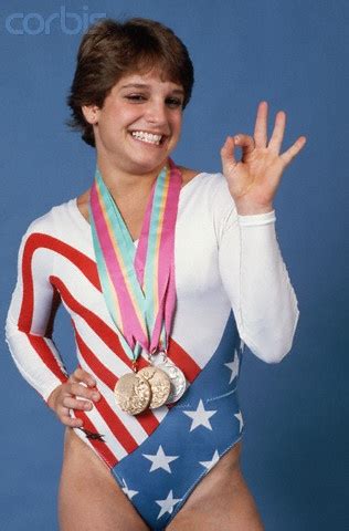 A Woman In A Bodysuit Holding Two Gold Medals And Making The Peace Sign With Her Hand
