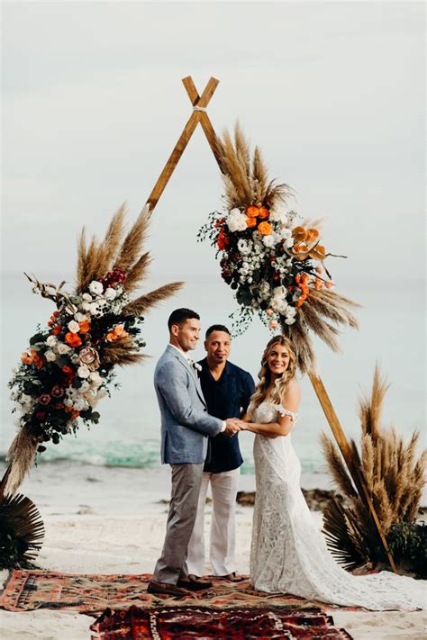 A Newly Married Couple Standing Under An Arch On The Beach With Flowers