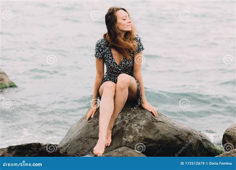 A Young Beautiful Girl In A Dress Is Sitting On Large Rocks Near The