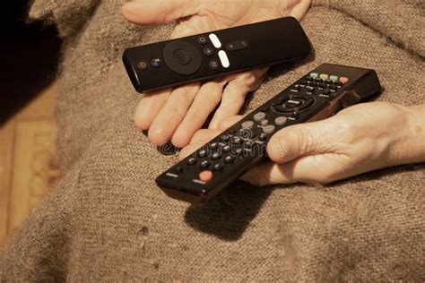 An Old Woman Holds In Her Hand Two Remotes From The Tv At Home Watching