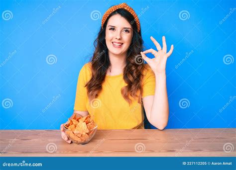 Young Beautiful Brunette Woman Sitting On The Table Eating Nachos Potato Chips Doing Ok Sign