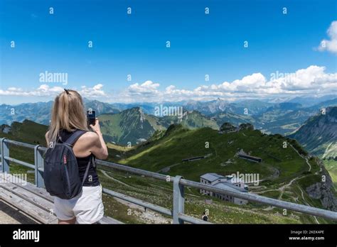Mature Adult Blonde Woman With Backpack Taking A Photo Of The Swiss Alps From A Lookout Point