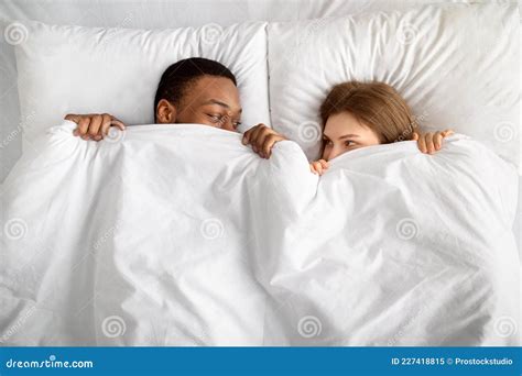 Top View Of Loving Interracial Couple Hiding Under White Blanket In Bed