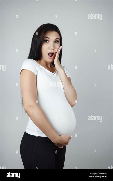 Beautiful Pregnant Brunette Woman Holding Her Pregnant Belly Looking Surprised On Isolated White