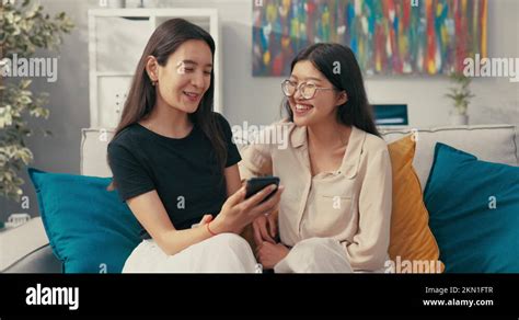 Two Attractive Asian Sisters Sit On Sofa In Room And Stare At Screen Of Their Stock Video