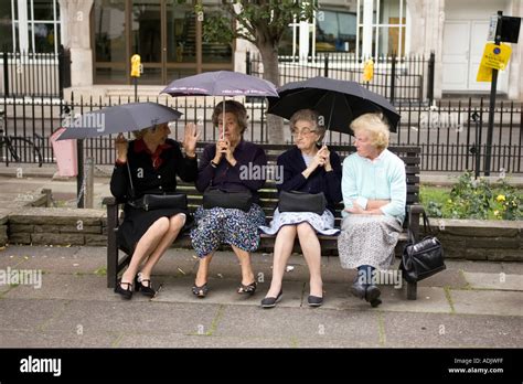 Old Women Sitting On A Bench Holding Umbrellas Under The Rain London Uk