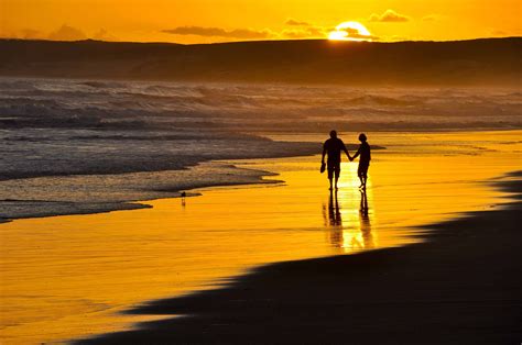 two people holding hands walking on the beach at sunset with waves coming in and setting sun