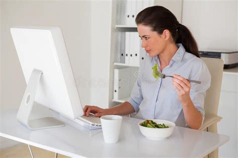 Casual Brunette Businesswoman Eating A Salad At Her Desk Stock Photo