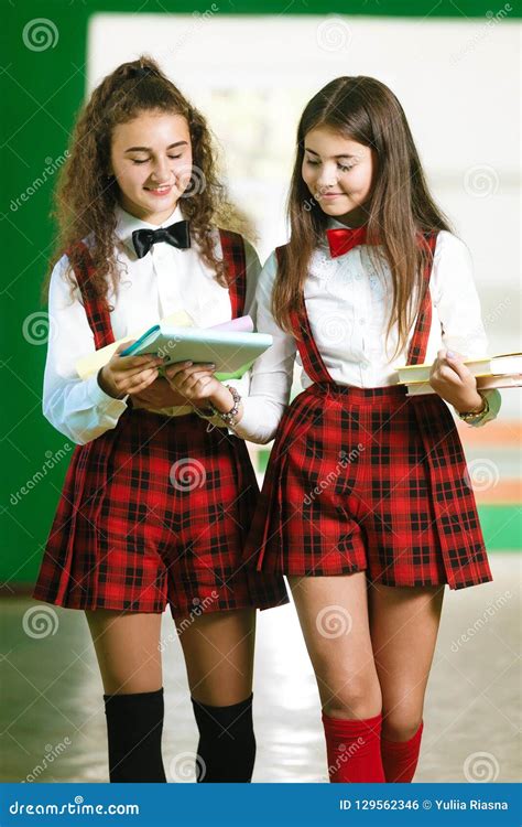 Two Schoolgirls In Red School Checkered Uniforms Stand In A Park Near A