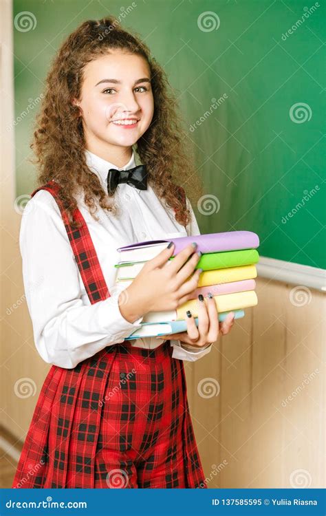Beautiful Schoolgirl Stands In The Classroom And Holds Books Against The Background Of The