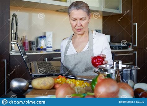 Mature Woman In The Kitchen Prepares Food And Uses Laptop Stock Photo