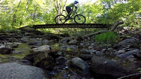A Man Riding A Bike Across A Wooden Bridge Over A Stream In The Woods