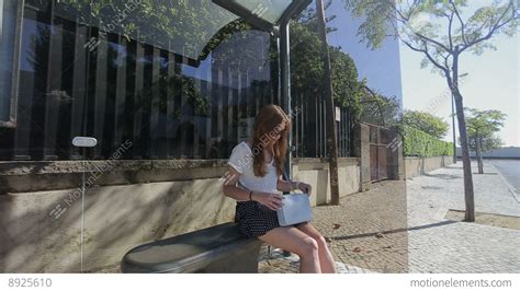 Young Woman Waiting And Talking By Phone At Glass Bus Stop In Skirt And