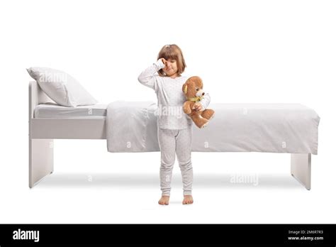 Girl In Pajamas Holding A Teddy Bear And Rubbing Eyes In Front Of A Bed