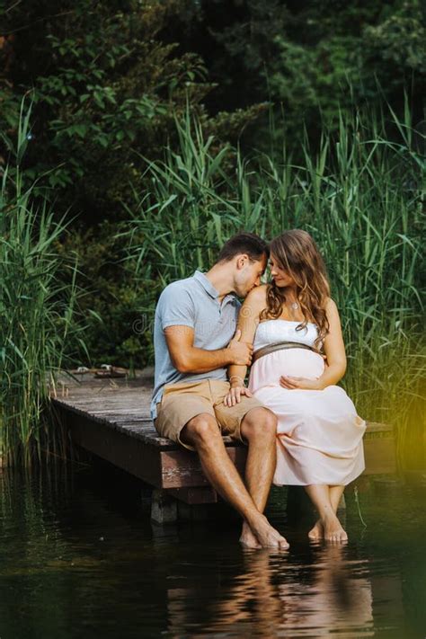 Pregnant Woman And Her Husband Sitting At A Lake While He Is Kissing