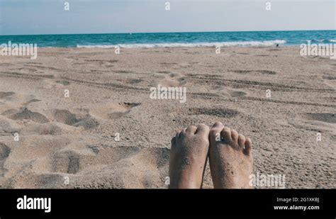 Female Feet Sunbathing On Beach Against The Background Of Sand And Blue