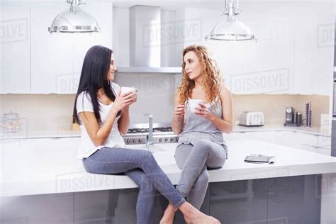 Two Young Women Sitting On Kitchen Counter Gossiping And Drinking