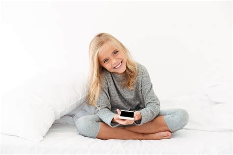 Fröhliches Mädchen Im Grauen Pyjama Das Smartphone Beim Sitzen Auf Dem Bett Hält Kostenlose Foto