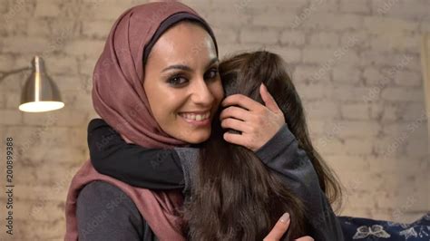 Young Beautiful Mother In Hijab Hugs Her Little Daughter Looks At Camera Sitting On Couch