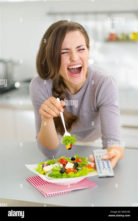 Laughing Young Woman Eating Salad And Watching Tv In Kitchen Stock
