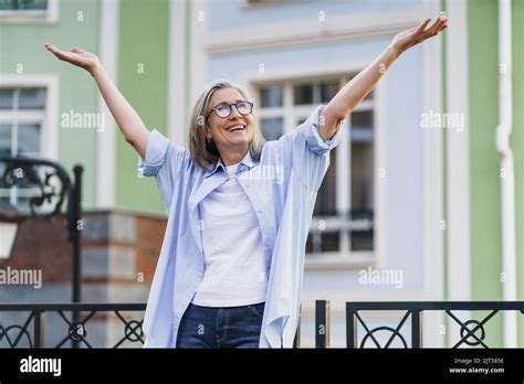 Happy Excited Mature Woman With Hands Lifted Up Wearing Glasses Standing Outdoors With Urban