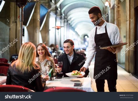 Positive Young Black Waiter Serving Terrace Restaurant Guests At Table