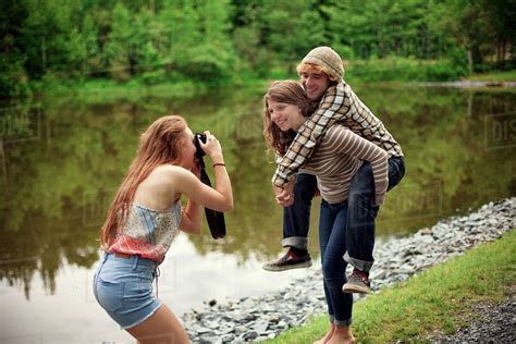 Girl Photographing Friend Carrying Boyfriend Piggyback Near Lake