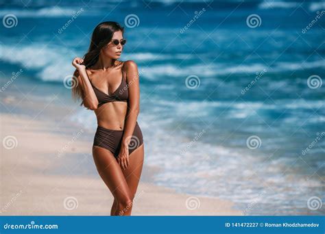 Tanned Model With Sunglasses In Brown Swimsuit Posing On White Sandy