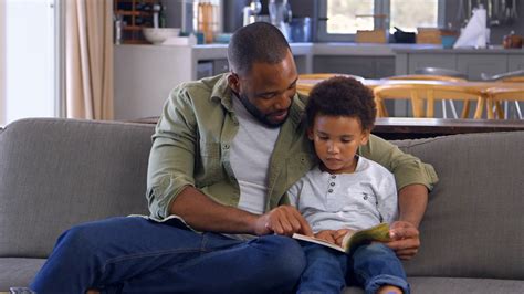 Father And Son Sitting On Sofa In Lounge Reading Book Together Stock