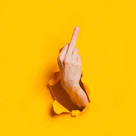 ジェスチャーの意味を示す女性の手は、黄色い紙の背景の穴からあなたをファックまたはファックオフ攻撃的なジェスチャー挑発の概念 プレミアム写真