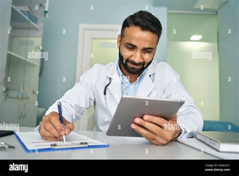 Indian Happy Male Doctor In Clinic Using Tablet Device Writing Health
