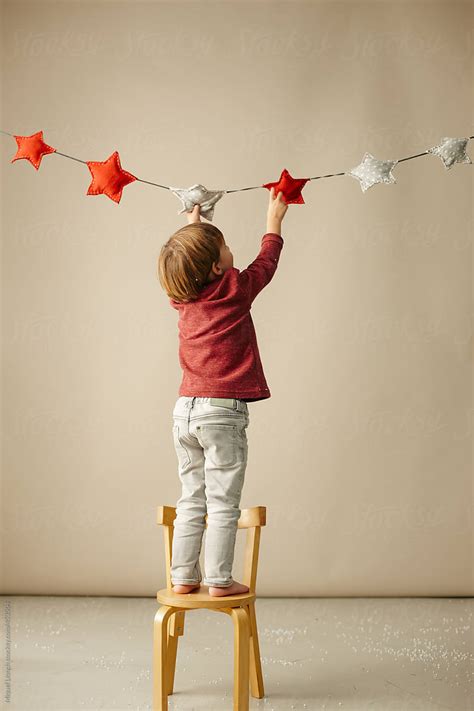Little Boy Standing On A Chair To Reach Hanging Christmas Stars By