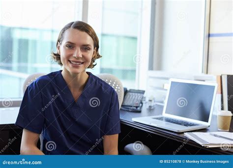 Portrait Of Nurse Wearing Scrubs Sitting At Desk In Office Stock Photo
